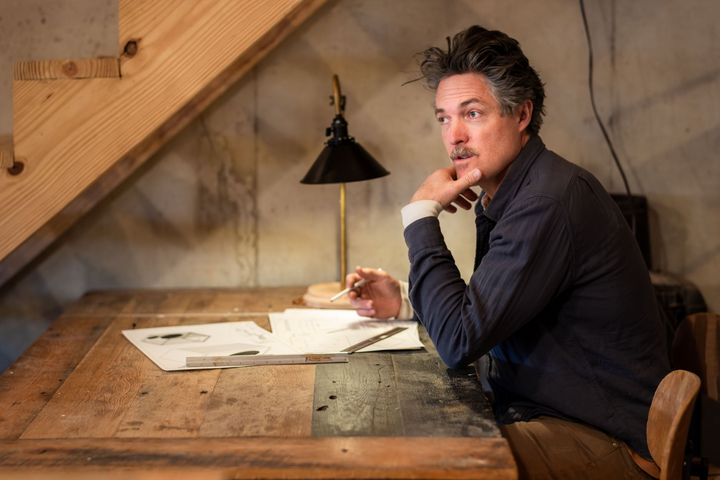Derek Sieg sitting at a wooden workbench in the Sieg workshop, chin on his hand, sketches on the table under a brass lamp.